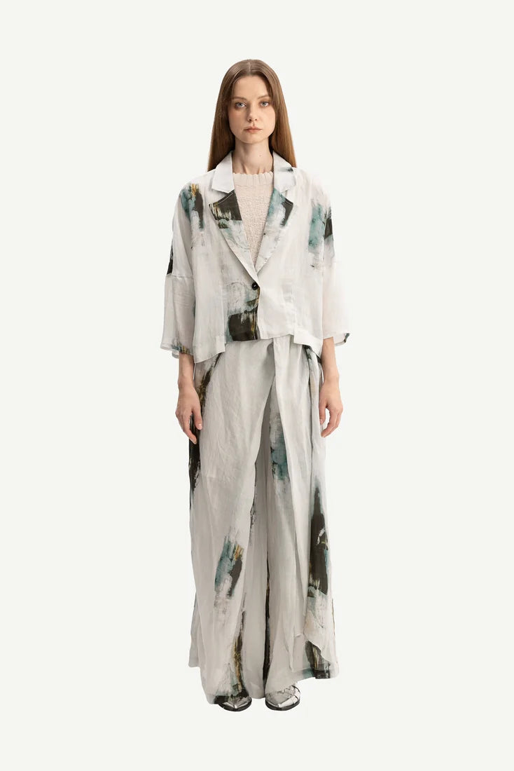 A model stands against a plain light gray studio background wearing a matching two-piece outfit consisting of a cropped, three-quarter sleeve jacket and wide-leg trousers, both in a light gray base with abstract watercolor-like green and black patterns.