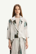 A woman modeling a light gray oversized blazer with abstract green and black watercolor-style prints, paired with matching light gray trousers and a textured cream top, against a solid off-white background.