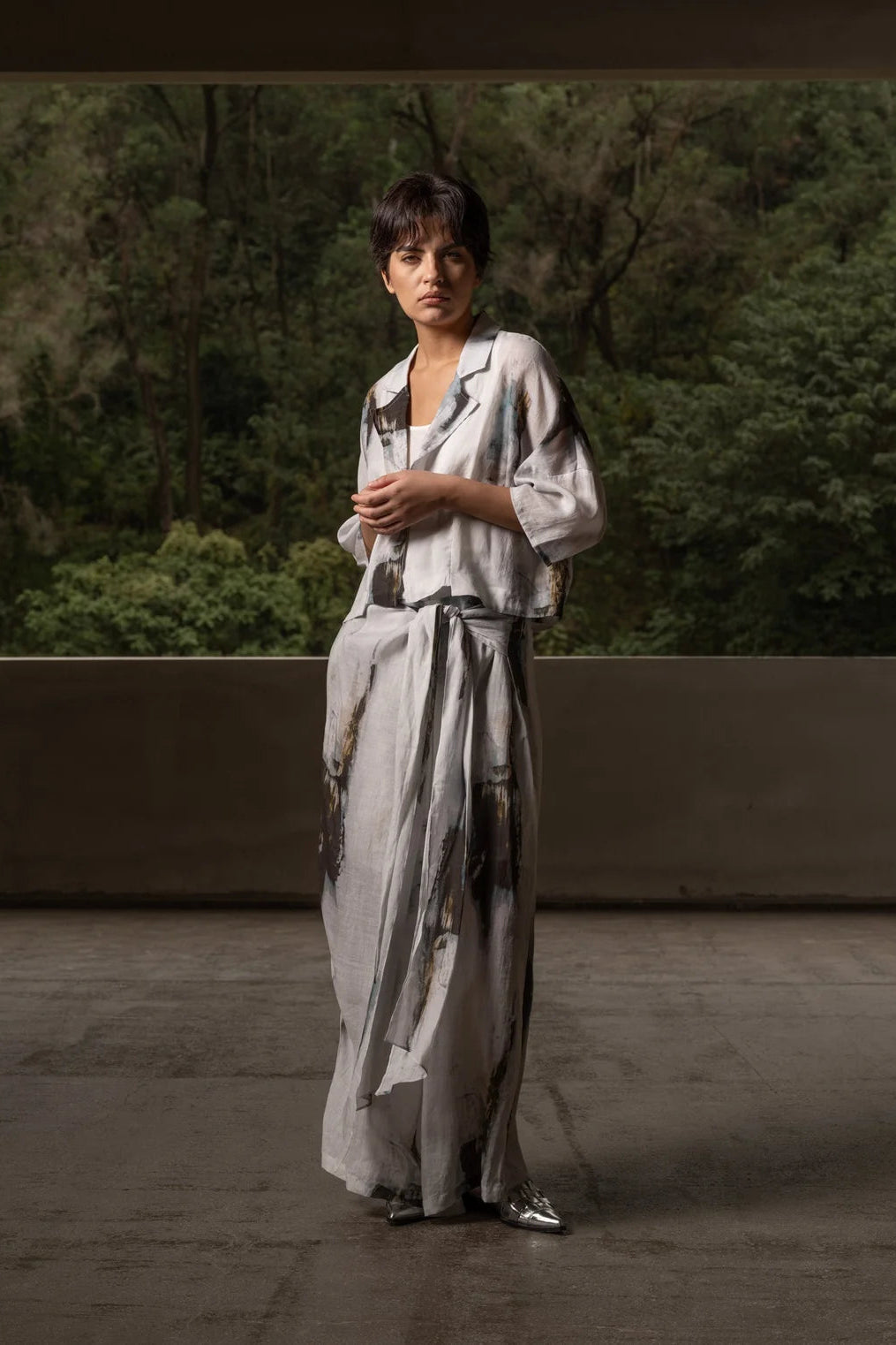 A model stands outdoors wearing a long, loose-fitting light gray dress with a dark abstract print, set against a background of green trees and a concrete wall.