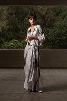 A model stands outdoors wearing a long, loose-fitting light gray dress with a dark abstract print, set against a background of green trees and a concrete wall.