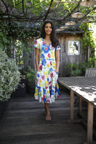 A woman stands barefoot on a wooden deck wearing a colorful floral midi dress with short sleeves and side pockets, set in a garden patio environment with a wooden table and pergola.