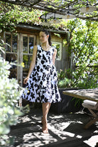 A woman wearing a sleeveless black and white floral print dress stands barefoot on a wooden deck in a garden setting with a rustic cabin and pergola in the background.