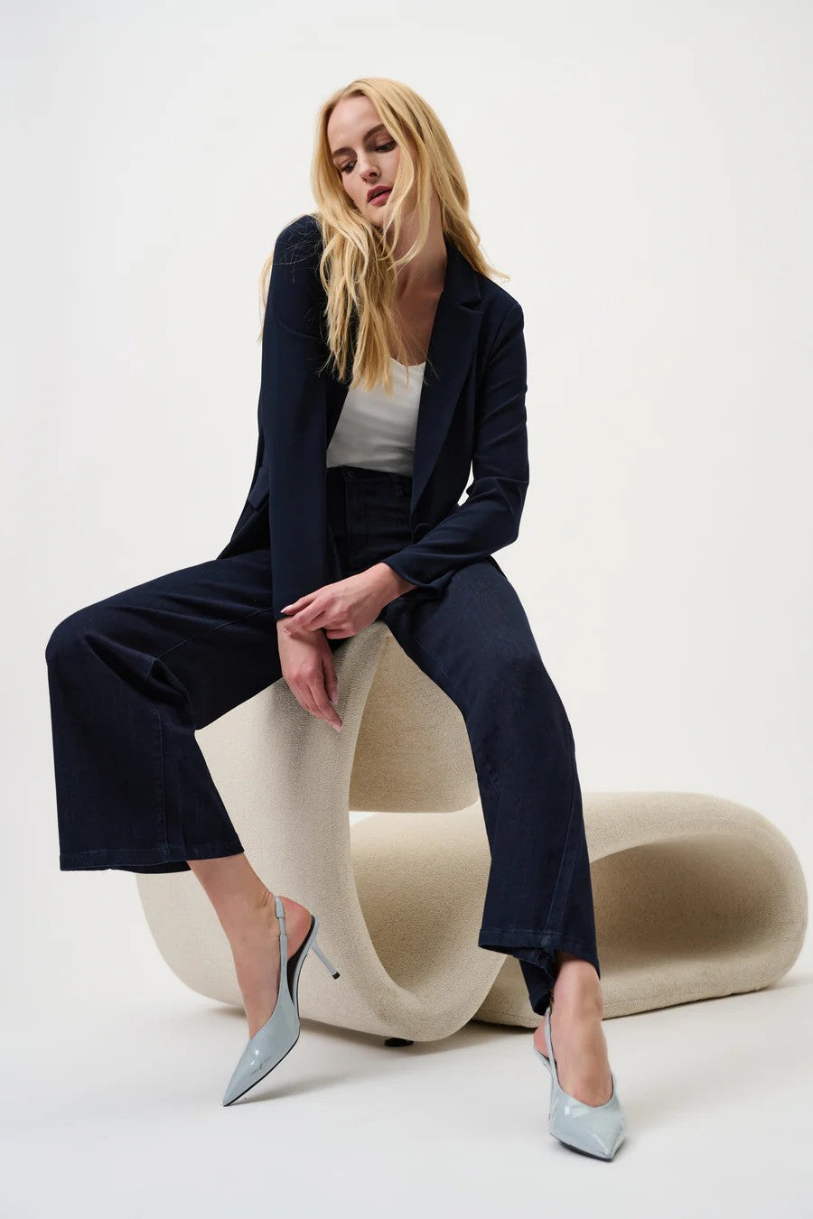 Woman wearing dark blue jacket and jeans with a white top sitting on a sculptural chair