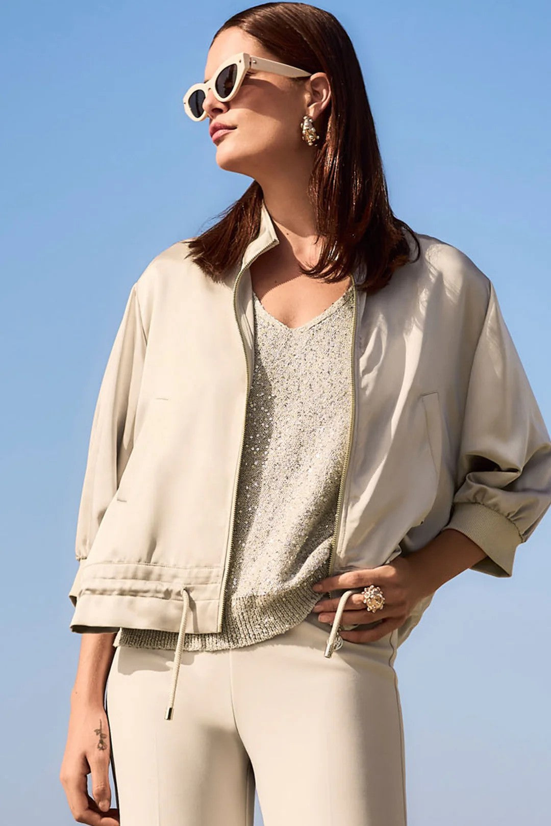 Woman wearing a beige jacket, sunglasses, and beige pants against a clear blue sky.