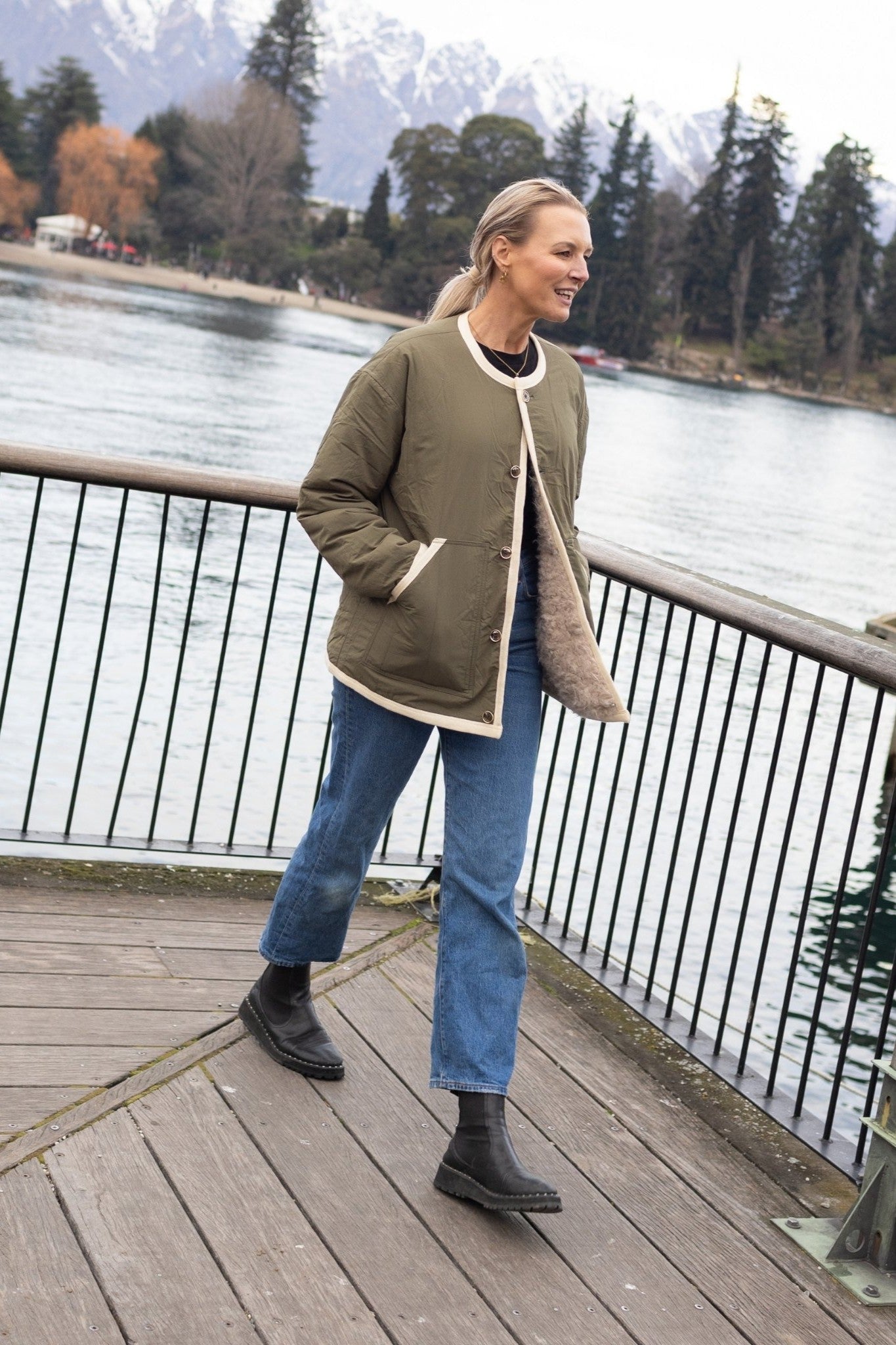 Person wearing olive jacket on a wooden deck by a lake with mountains in the background