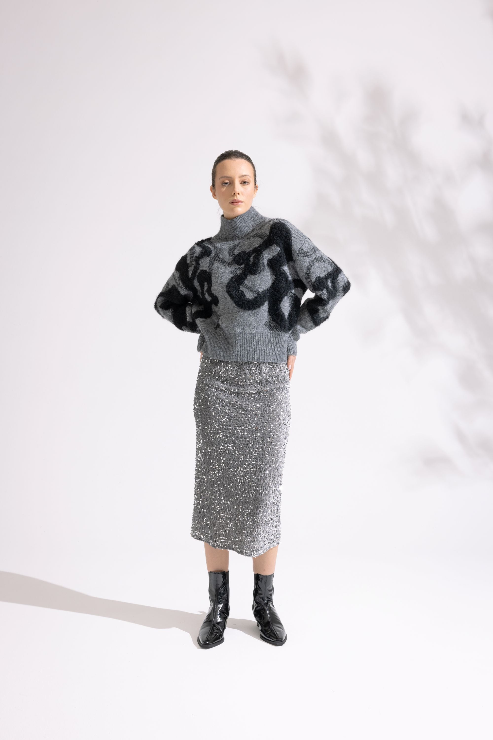 Person wearing a gray turtleneck sweater with black abstract patterns and a patterned skirt on a white background