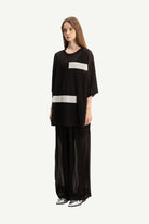 A model wearing a black wide-sleeve top with two horizontal white stripes and matching black wide-leg sheer pants, standing against a solid off-white background.