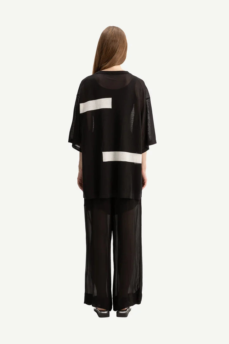 A model viewed from the back wearing a black sheer mesh two-piece outfit with wide-leg pants and a short-sleeve top featuring horizontal white stripe accents.