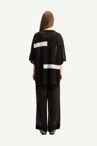 A model viewed from the back wearing a black sheer mesh two-piece outfit with wide-leg pants and a short-sleeve top featuring horizontal white stripe accents.