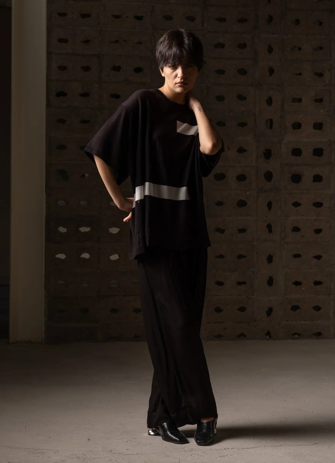 A model wearing a black oversized short-sleeve top with a silver reflective stripe and matching black wide-leg trousers, standing in front of a textured concrete wall.