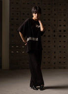 A model wearing a black oversized short-sleeve top with a silver reflective stripe and matching black wide-leg trousers, standing in front of a textured concrete wall.