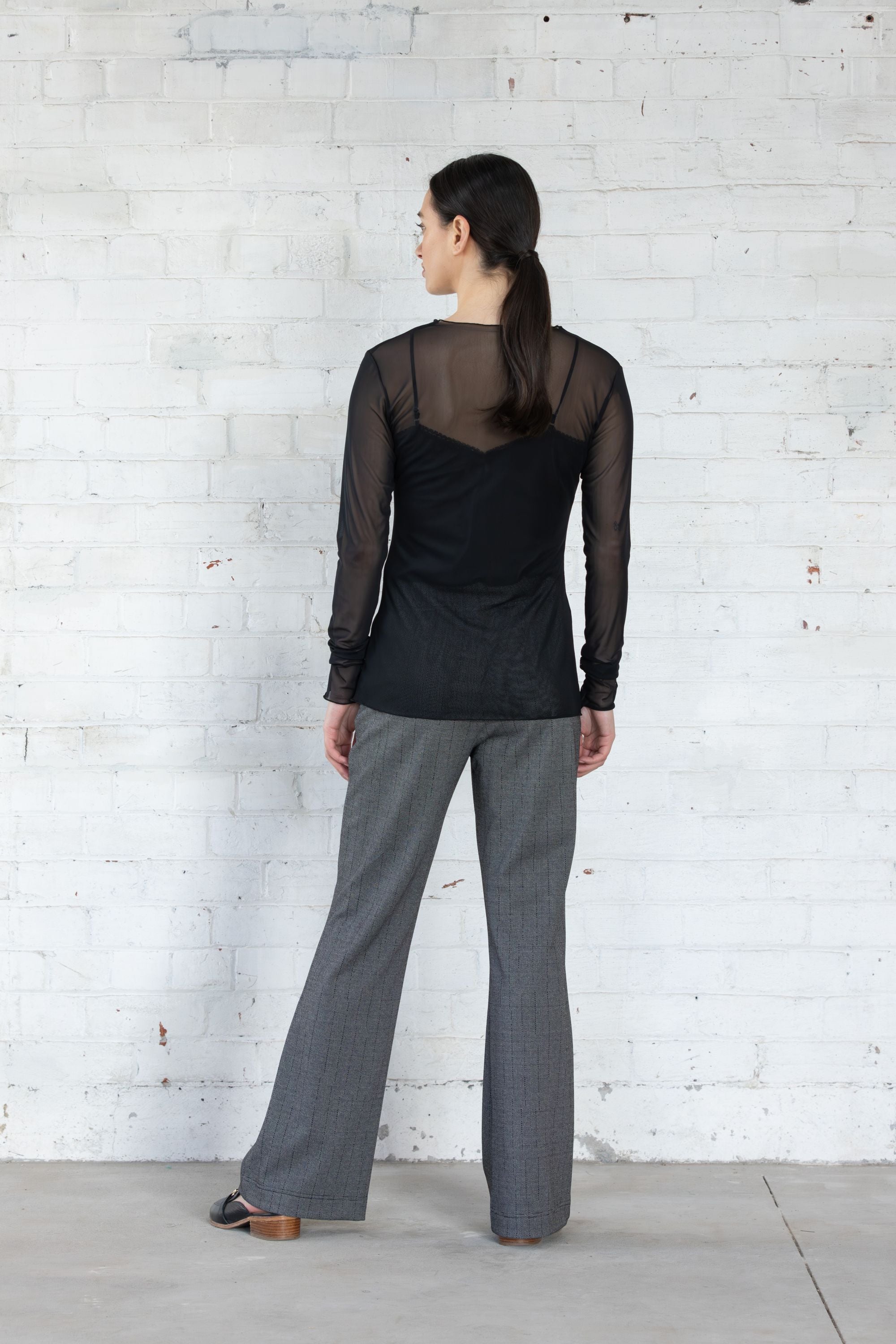 Person wearing a black sheer top and gray pants against a white brick wall back view