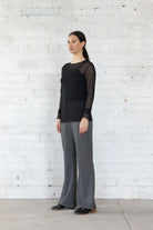 Woman wearing a black top and gray pants standing against a white brick wall.