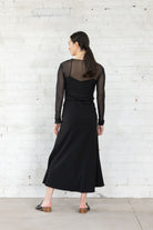 Woman wearing a black dress with sheer sleeves against a white brick wall back view