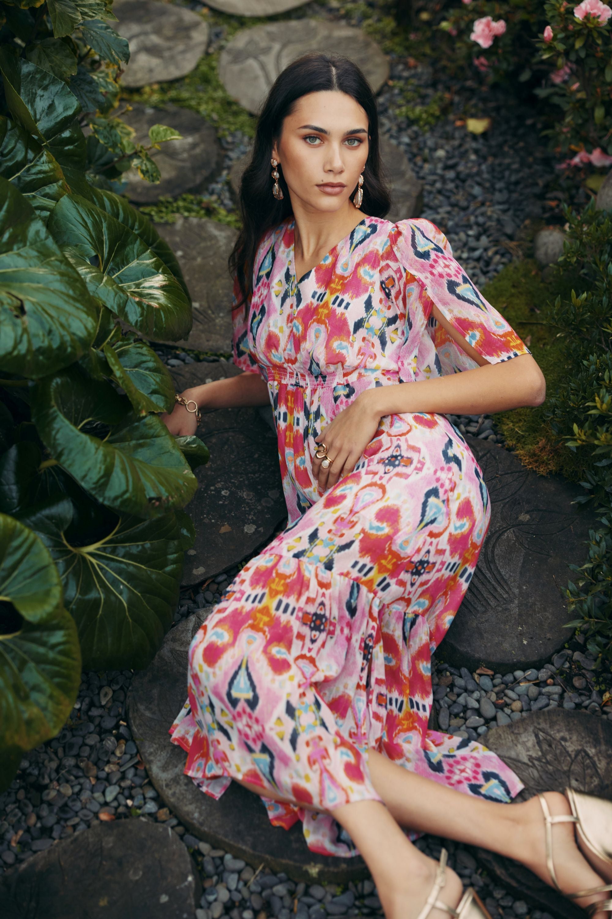 Woman in a colorful floral dress sitting on a stone path surrounded by greenery.