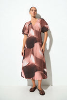 A woman modeling a midi-length dress with a pink, brown, and beige abstract floral pattern, featuring short puff sleeves and a V-neckline, set against a plain white studio background.