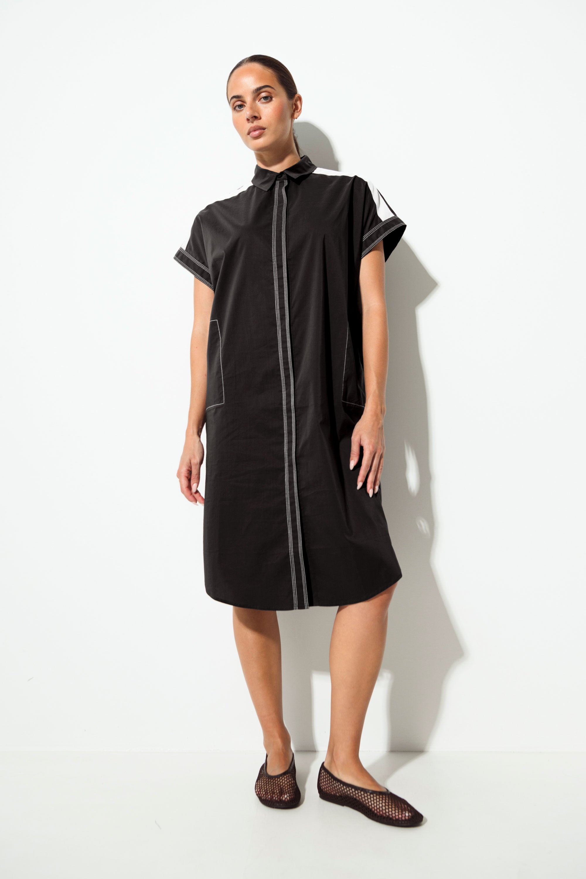A woman modeling a black short-sleeve midi dress with contrasting light gray piping details, standing against a white studio background.