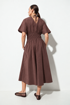 A woman wearing a brown short-sleeve midi dress with a smocked waist and tie-back neckline, shown from the back against a white studio background.