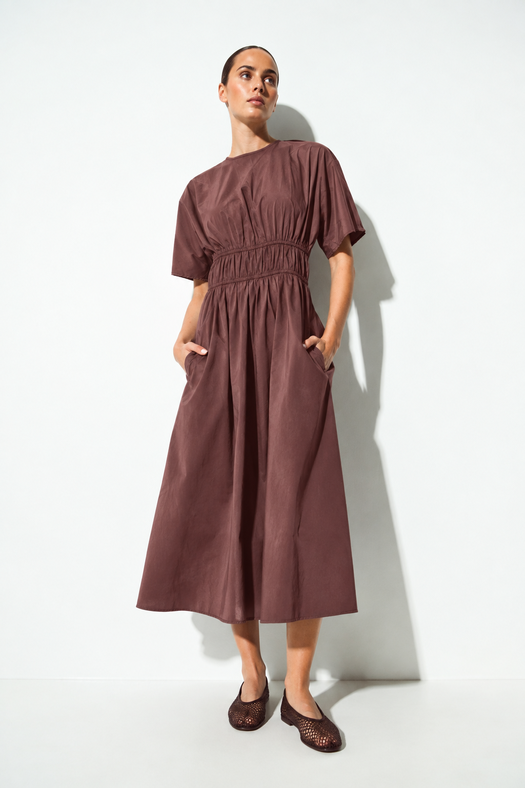A woman modeling a short-sleeve midi dress in a dusty plum-brown color, featuring a smocked waist and side pockets, standing against a white wall with a soft shadow.