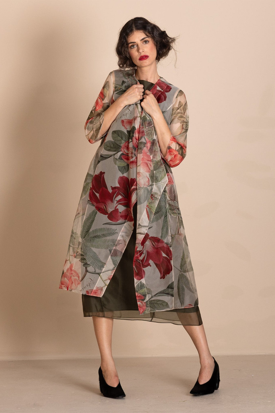 A woman models a long, sheer, open-front duster coat featuring a large red and pink floral print on a gray background. She is wearing a black midi dress underneath and black pointed-toe heels, standing against a solid beige studio backdrop.