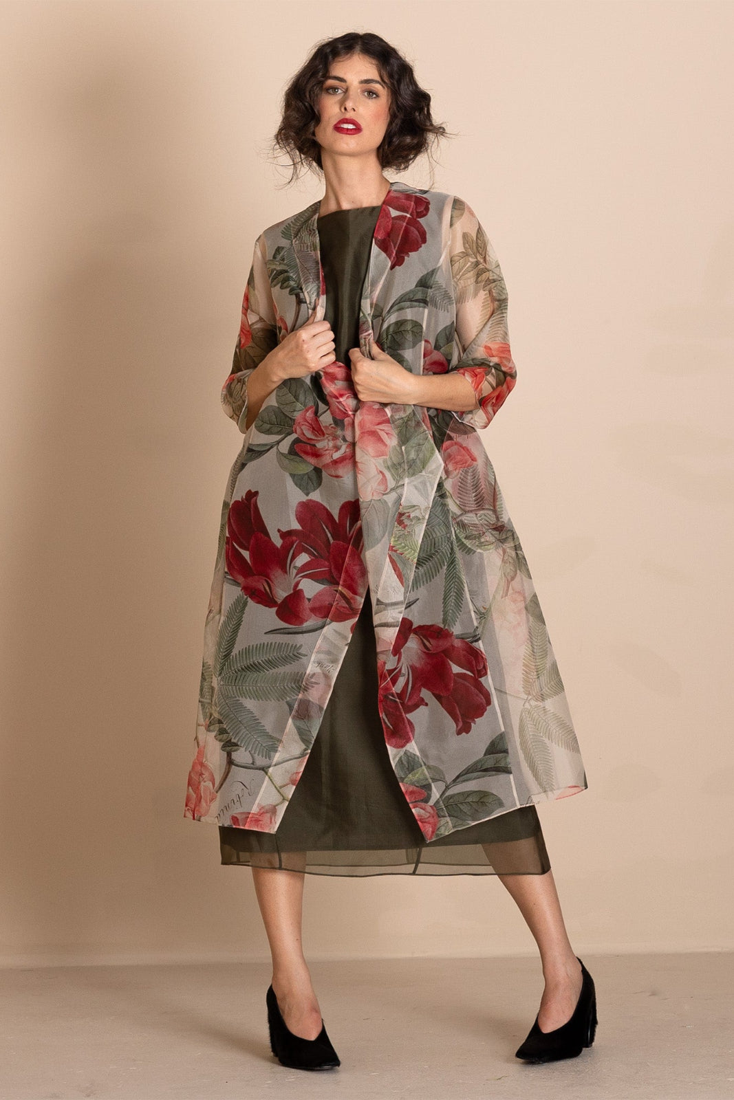 A woman models a sheer, lightweight open-front duster jacket featuring a large red and green floral print over a dark olive green dress, set against a solid beige studio background.