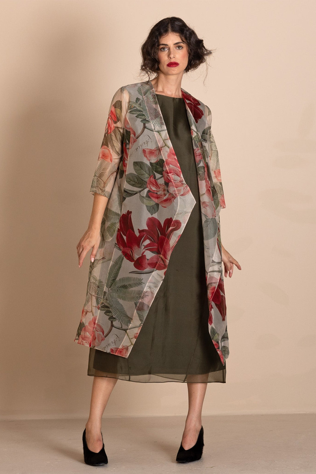 A woman models a sheer, open-front midi duster coat featuring a red and green floral print over a dark olive green dress, set against a plain beige studio background.