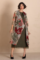 A woman models a sheer, open-front midi duster coat featuring a red and green floral print over a dark olive green dress, set against a plain beige studio background.