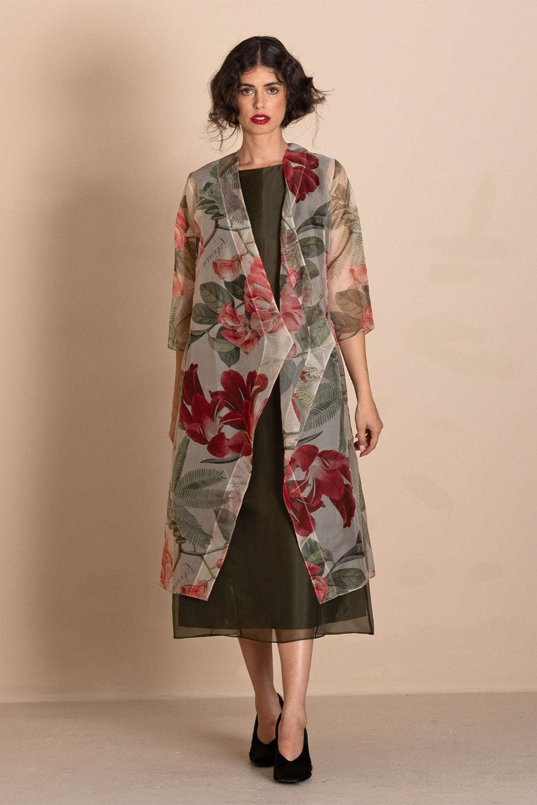 A woman models a sheer floral kimono jacket over an olive green dress, standing against a solid beige studio background.