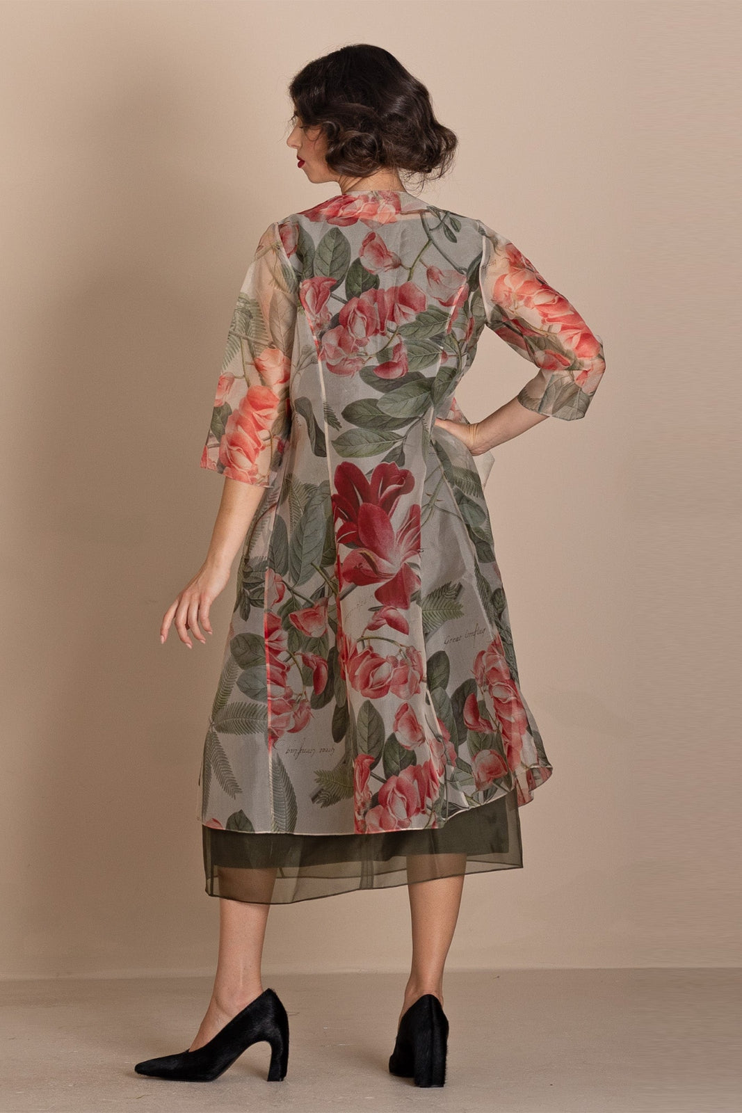 A woman is shown from the back wearing a sheer floral printed tunic dress with three-quarter sleeves, featuring red and pink flowers with green leaves on a gray base, paired with black heels, set against a solid beige studio background.