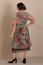 A woman is shown from the back wearing a sheer floral printed tunic dress with three-quarter sleeves, featuring red and pink flowers with green leaves on a gray base, paired with black heels, set against a solid beige studio background.
