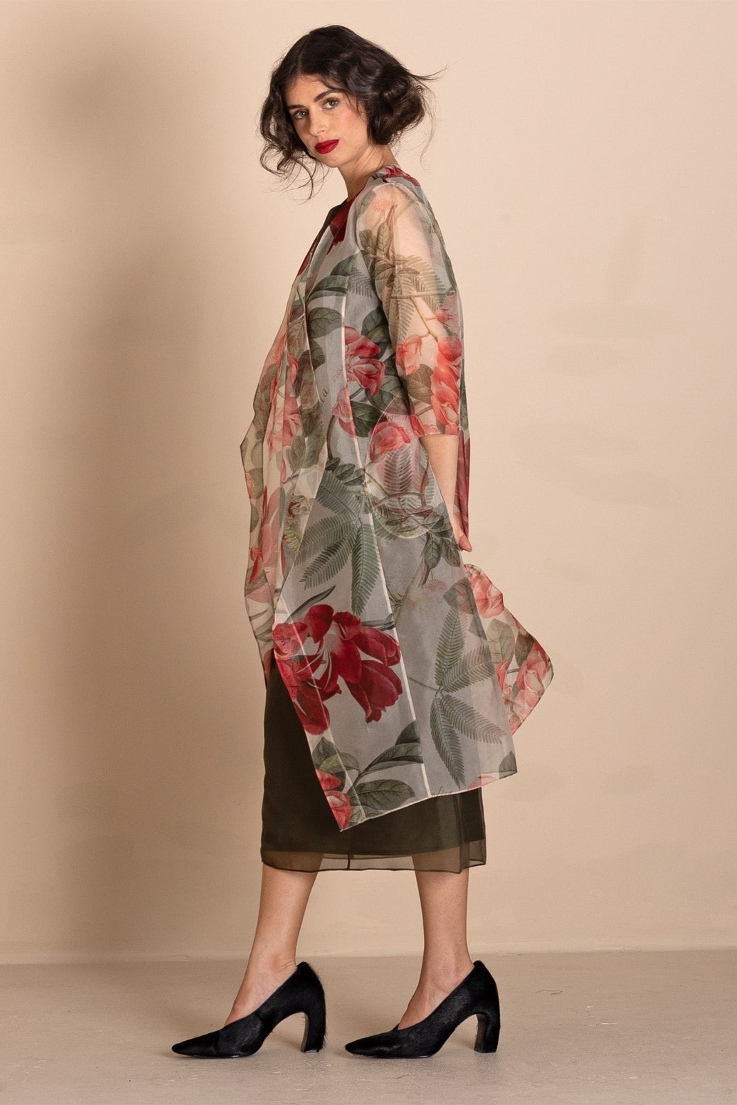 A woman models a sheer, lightweight open-front cardigan featuring a vibrant red and green floral print over a dark olive green midi dress, styled with black heels against a solid beige studio backdrop.