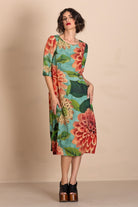 A woman models a knee-length dress with a vibrant floral print featuring large red and orange flowers, green leaves, and a turquoise blue background. She is wearing black platform heels against a solid beige studio backdrop.