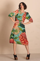 A woman models a knee-length dress with a vibrant floral print featuring red and orange flowers, green leaves, and a turquoise blue background. She is posing with hands on hips against a solid beige studio backdrop.