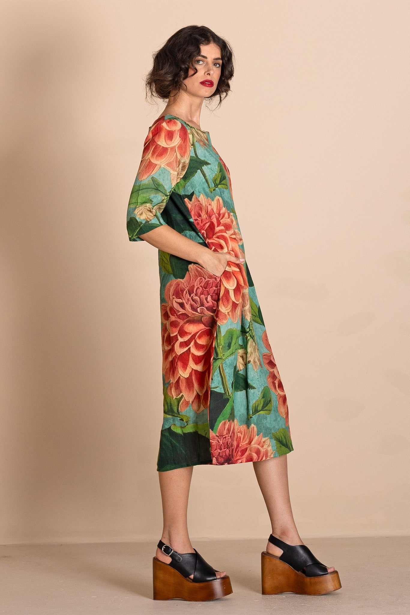 A woman models a knee-length dress with a vibrant green base and a large red and orange floral print, styled with black platform sandals against a solid beige studio background.