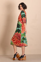 A woman modeling a knee-length A-line dress with a vibrant floral print featuring red, orange, and green dahlias on a beige background.