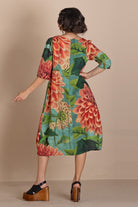 A woman stands with her back to the camera wearing a knee-length dress with a large floral print in red, orange, green, and teal, paired with black wedge sandals, against a solid beige studio background.