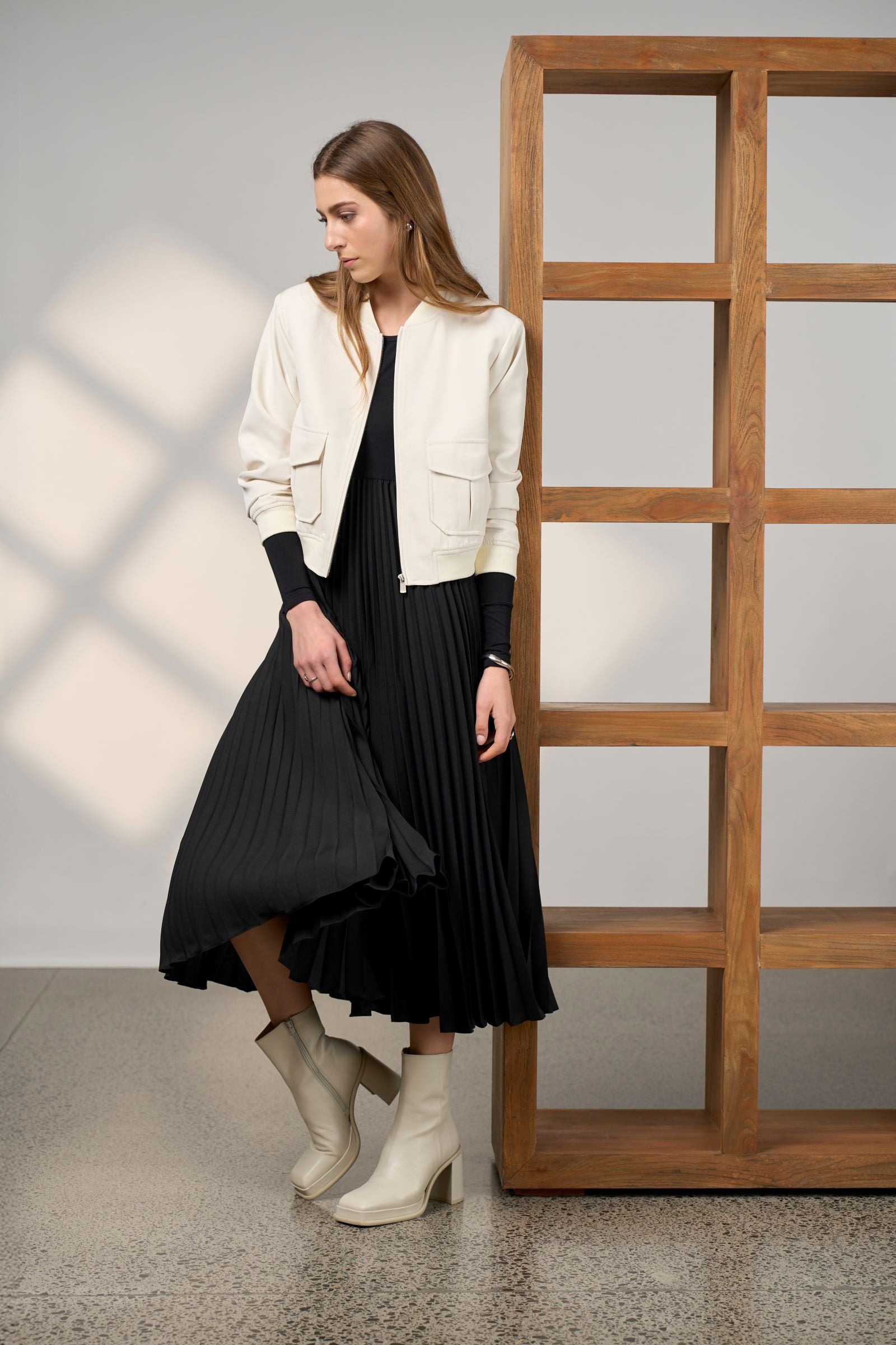 Woman in a black pleated dress and white jacket standing in a room with wooden shelves.