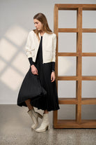 Woman in a black pleated dress and white jacket standing in a room with wooden shelves.
