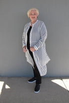 An older woman with short blonde hair stands outdoors against a gray wall, wearing a long-sleeve, open-front striped cardigan over a black top and black pants with black sneakers.