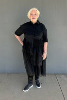 An older woman with short blonde hair stands against a gray concrete wall wearing a black short-sleeve button-up shirt with a gathered waist, layered sheer black tulle skirt, black baggy pants, and black sneakers with white soles.