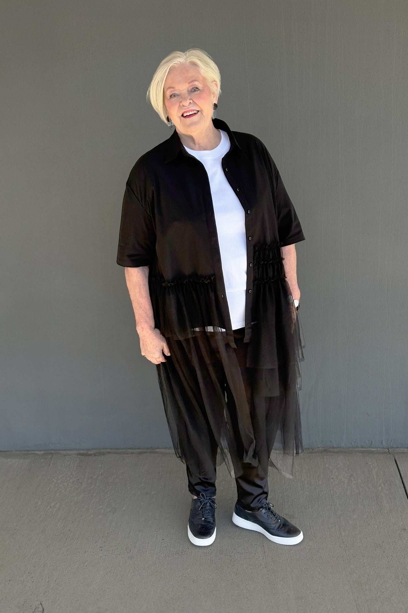 An older woman with short blonde hair stands against a plain gray wall wearing a black sheer tulle shirt-jacket, loose black pants, a white t-shirt, and black sneakers with white soles.