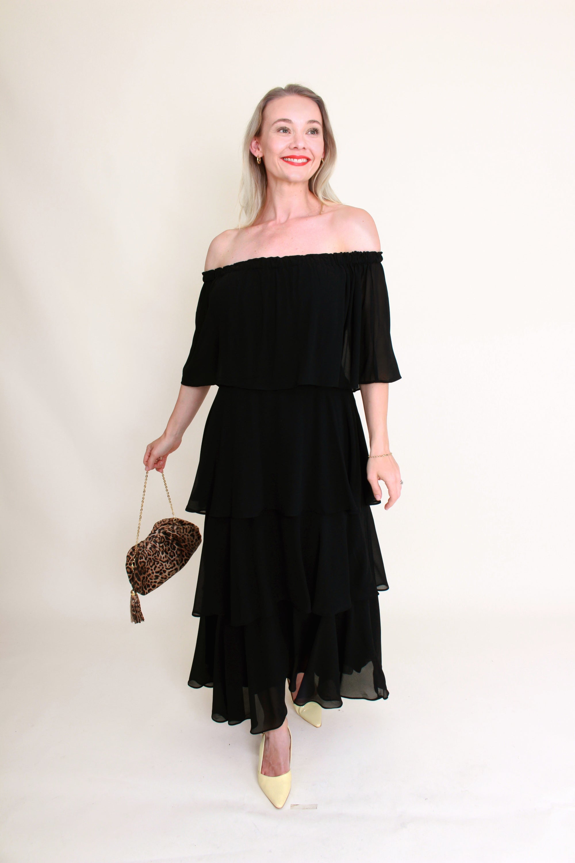 Woman wearing a black off-shoulder dress holding a small bag against a plain background