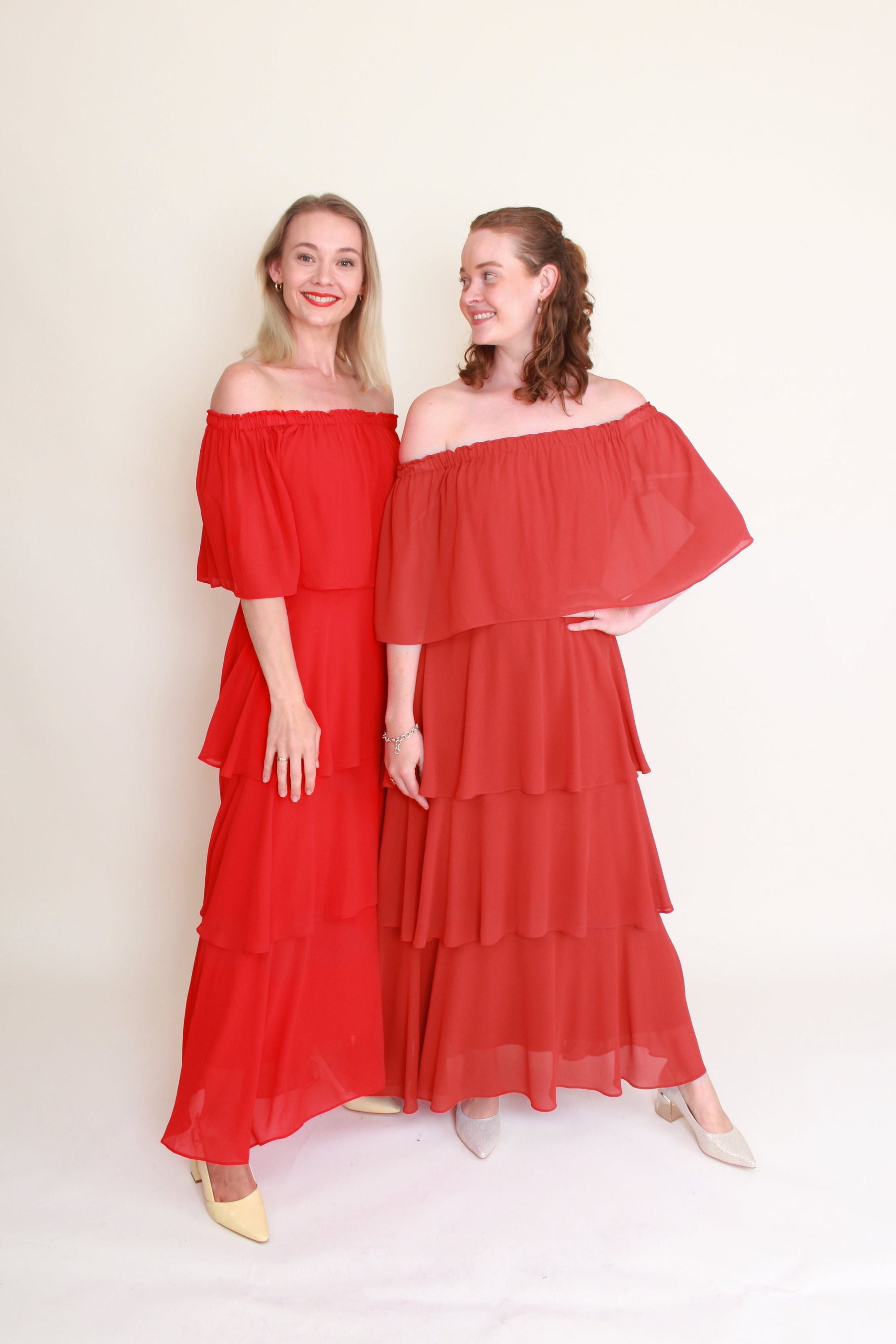 Two women wearing red and terracottaoff-shoulder dresses against a white background