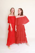 Two women wearing red and terracottaoff-shoulder dresses against a white background