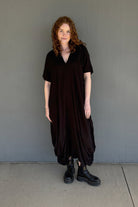A woman with curly light brown hair stands against a gray concrete wall wearing a loose, oversized short-sleeve black dress and black leather boots.