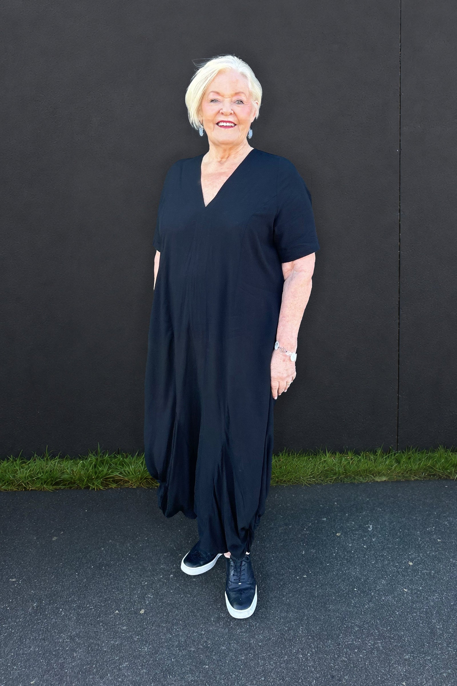 An older woman with short white hair stands outdoors against a dark wall, wearing a loose black V-neck maxi dress with short sleeves and black sneakers.