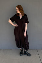 A woman with curly red hair models a loose, dark brown short-sleeve midi dress with a V-neckline, paired with black combat boots, standing against a plain gray wall.