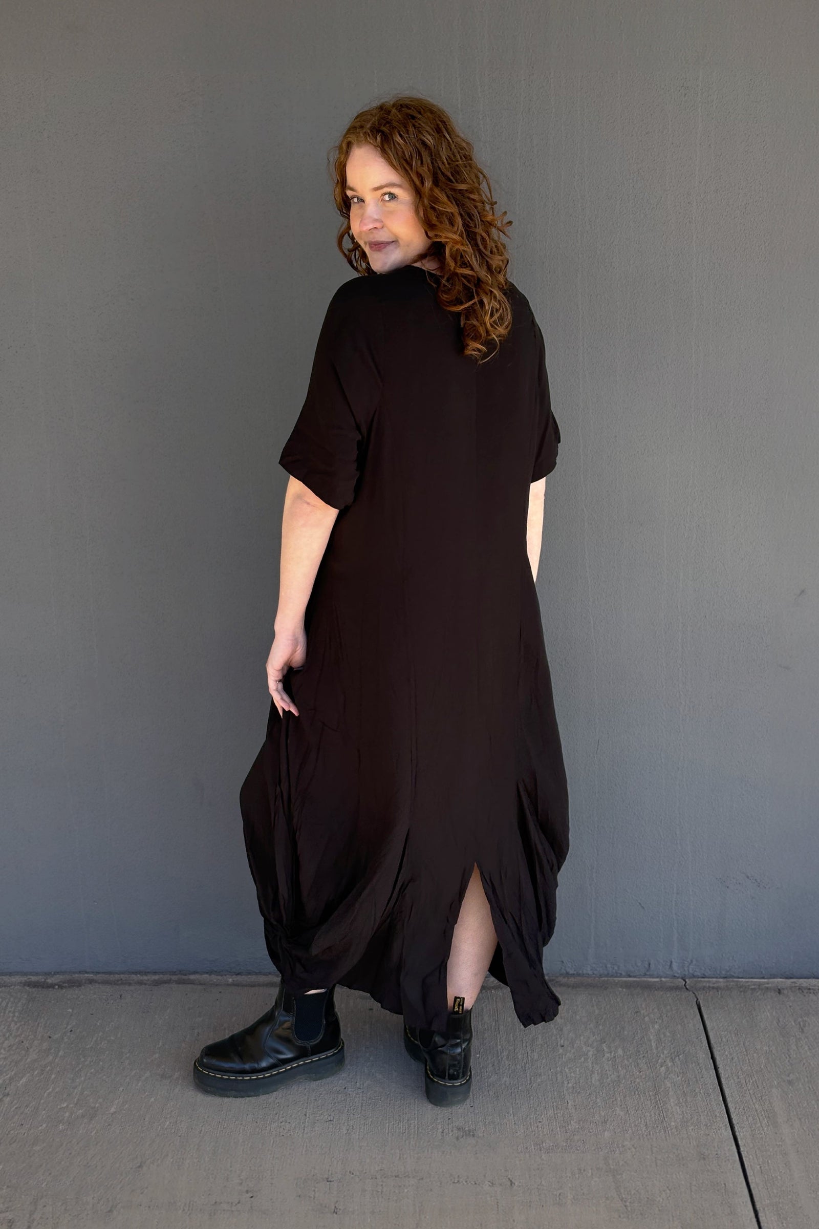 A woman with curly brown hair is shown from the back wearing a loose, oversized black dress with short sleeves and black boots, standing against a plain gray wall.