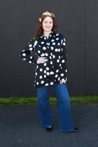 A woman with curly brown hair stands outdoors against a dark wall, wearing a black long-sleeve button-up shirt with large white polka dots and blue wide-leg jeans.