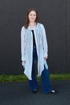 A woman stands outdoors against a dark gray wall wearing a long-sleeve white shirt with thin vertical black stripes, a black top underneath, wide-leg blue jeans, and black flats.